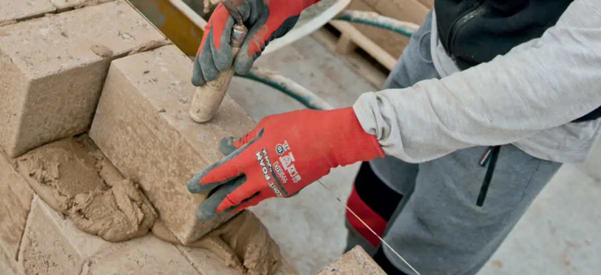 Operario con guantes rojos asentando un bloque de tierra sobre una capa de mortero en un muro en construcción.