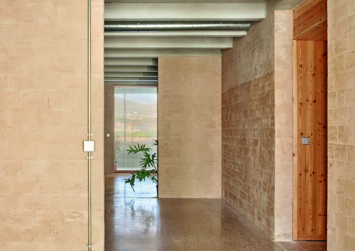 Pasillo interior con muros de bloques de tierra de tono arena, techo de vigas de hormigón y puerta de madera.