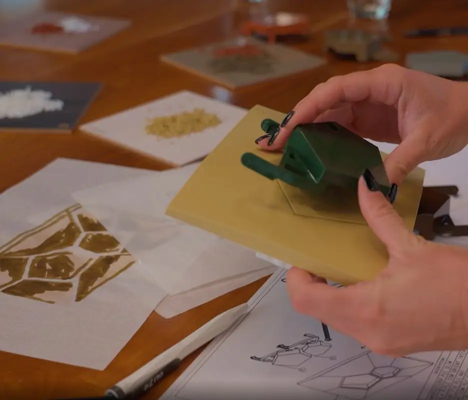 Manos que sostienen un prototipo de pulsador sobre una mesa con bocetos del patrón facetado y muestras de material