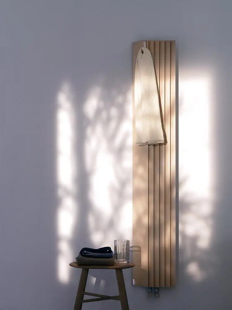 Radiador vertical de aluminio con módulos plisados en color arena, con una toalla colgada y un taburete de madera