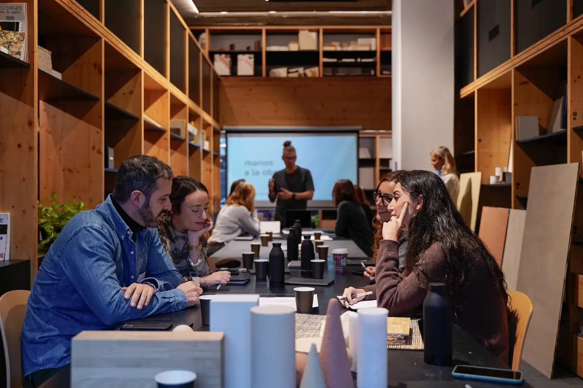 Participantes sentados a una mesa larga con muestras de materiales mientras un ponente presenta ante una pantalla