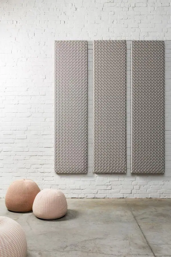 Tres paneles acústicos textiles grises colgados en vertical sobre una pared de ladrillo blanco, con pufs de punto en el suelo