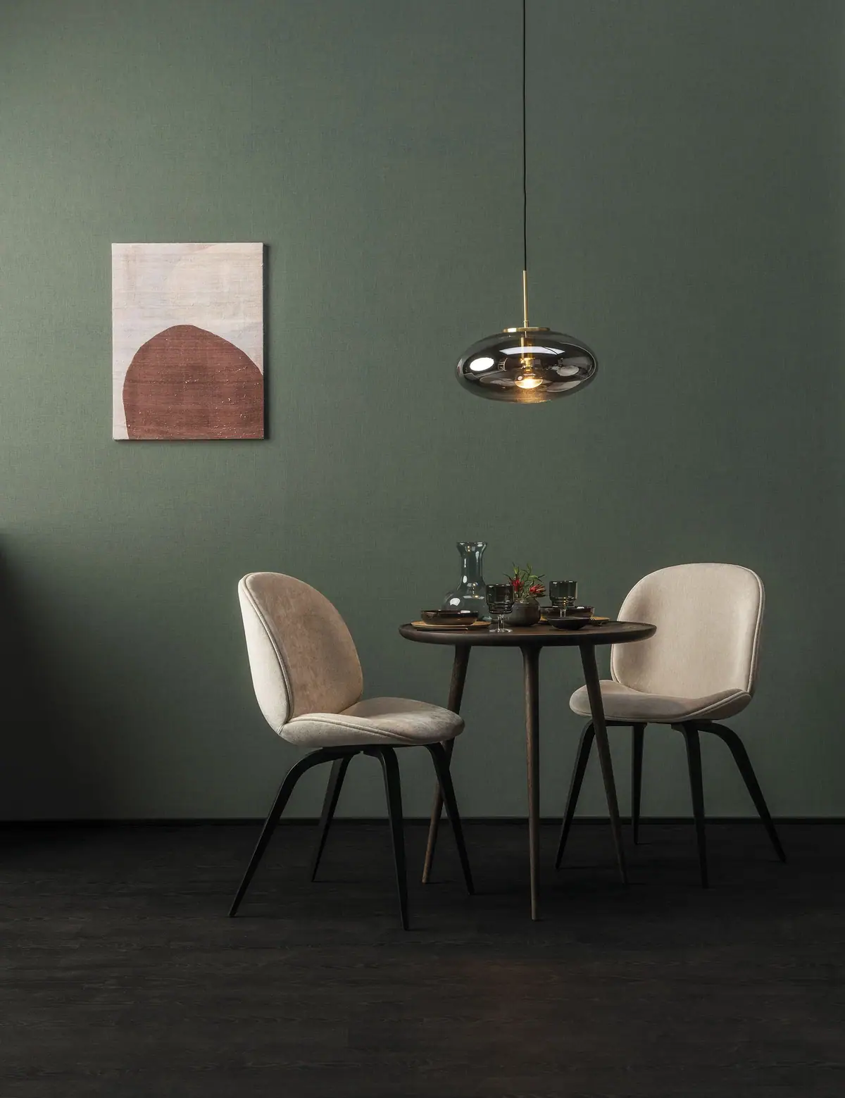 Comedor con dos sillas tapizadas, mesa redonda de madera y lámpara de vidrio ante una pared revestida en verde musgo.