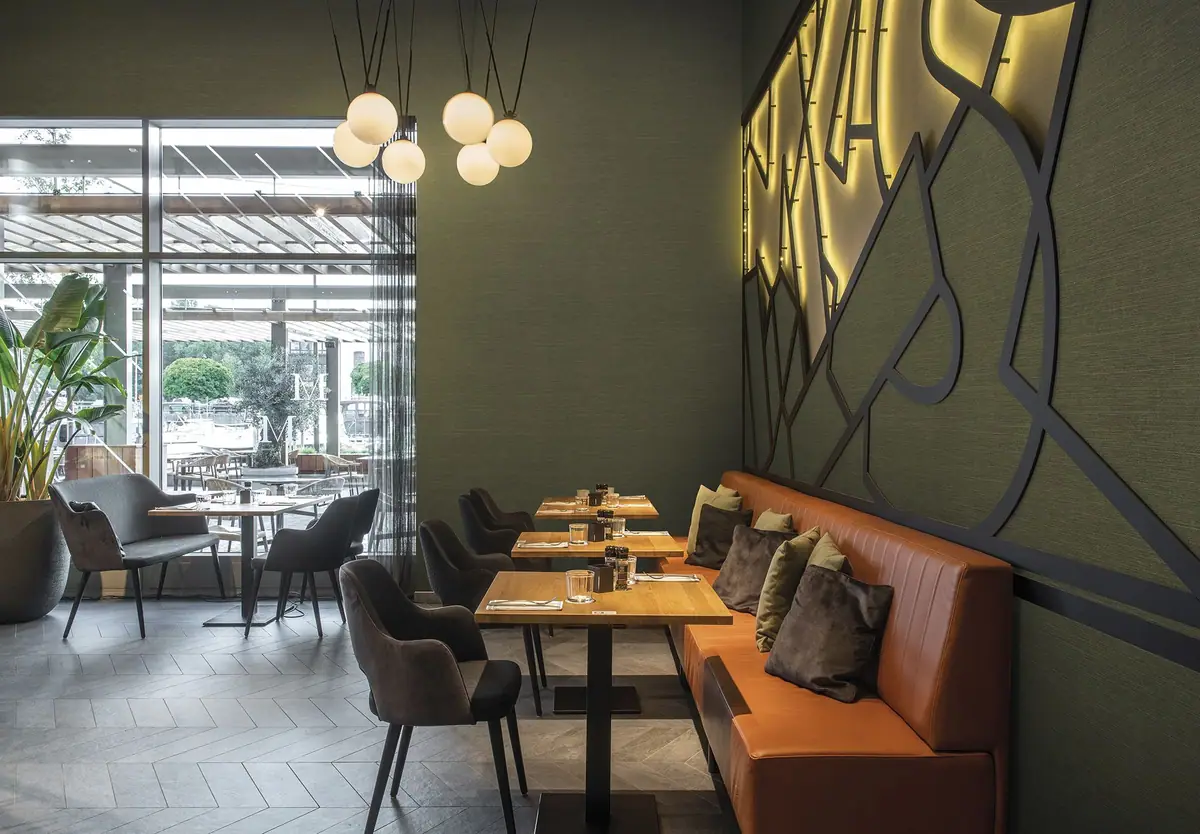 Restaurante con bancada de piel color teja, mesas de madera y paredes verde oscuro con un mural geométrico iluminado.