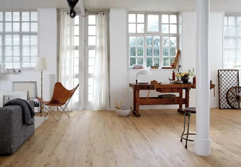 Salón diáfano con grandes ventanales, pavimento de madera clara, banco de carpintero y butaca de cuero