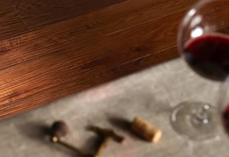 Pavimento de madera de tonos cálidos junto a un suelo gris, con una copa de vino tinto, un sacacorchos y un tapón de corcho