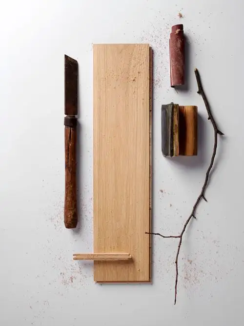 Tabla de roble claro sobre fondo blanco, con una cuchilla de mango de madera, astillas y una rama