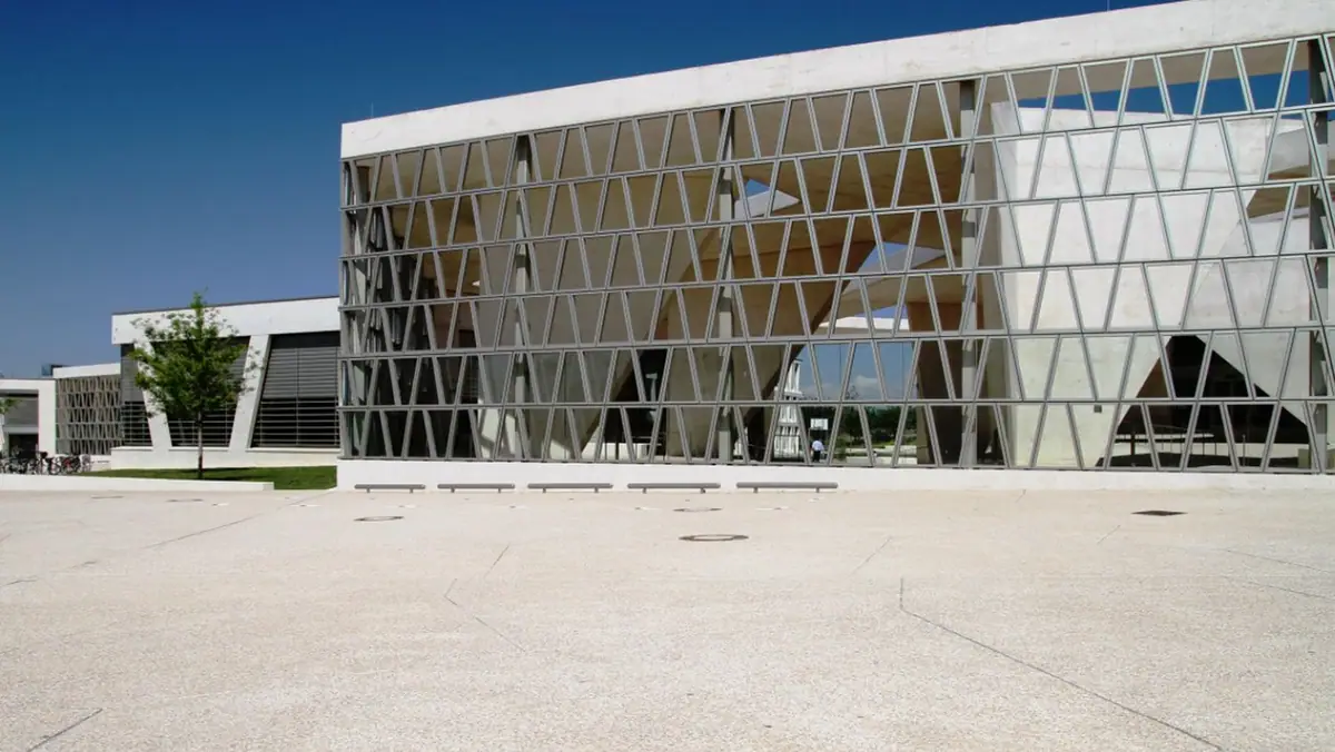 Explanada de hormigón claro con árido visto frente a un edificio con celosía metálica de piezas trapezoidales