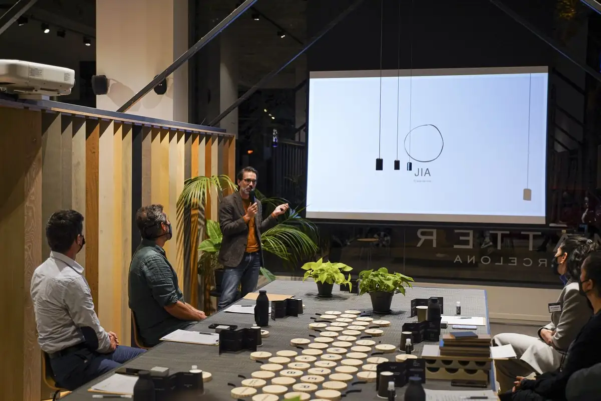 Un ponente habla ante la proyección del logotipo de las casas JIA, junto a la mesa con muestras de materiales