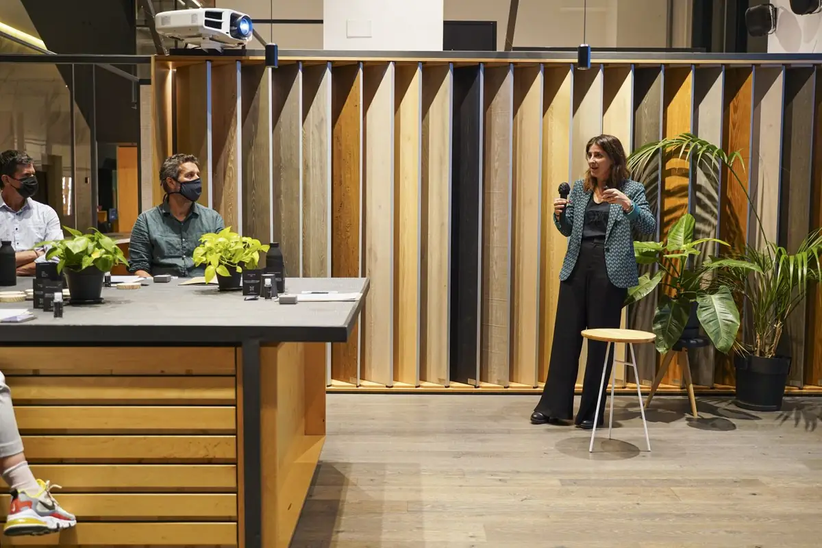 Una ponente de pie con micrófono entre plantas, junto a la mesa de los ponentes