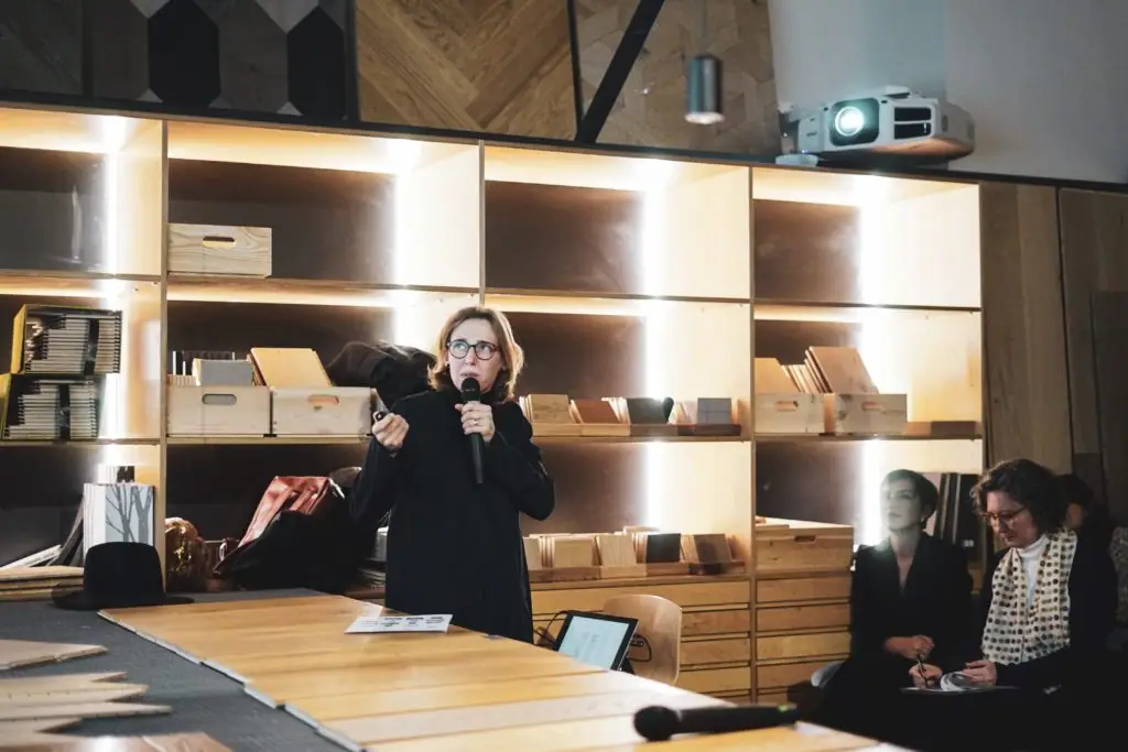 Una de las arquitectas invitadas habla con micrófono junto a la mesa de materiales mientras otras dos siguen la charla sentadas