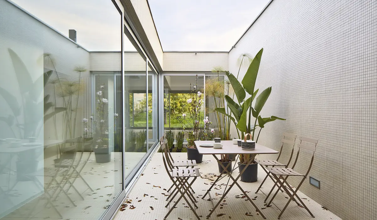 Patio interior con suelo de mosaico blanco, mesa con sillas plegables y plantas, rodeado de carpinterías de vidrio