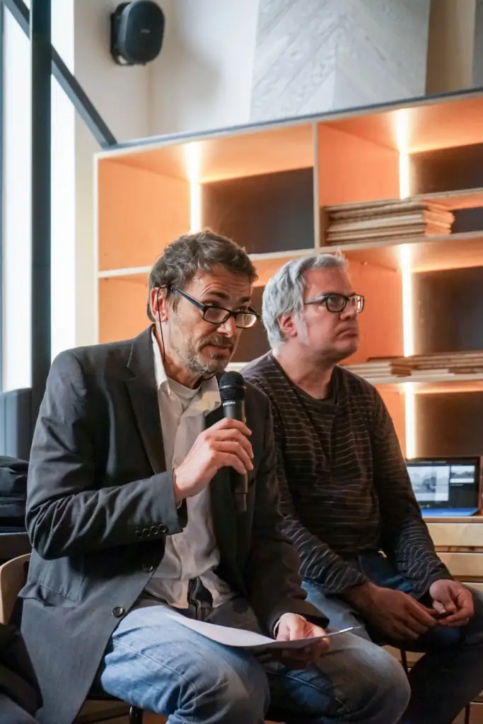 Ponente con gafas hablando por micrófono, sentado junto a otro asistente ante una estantería iluminada