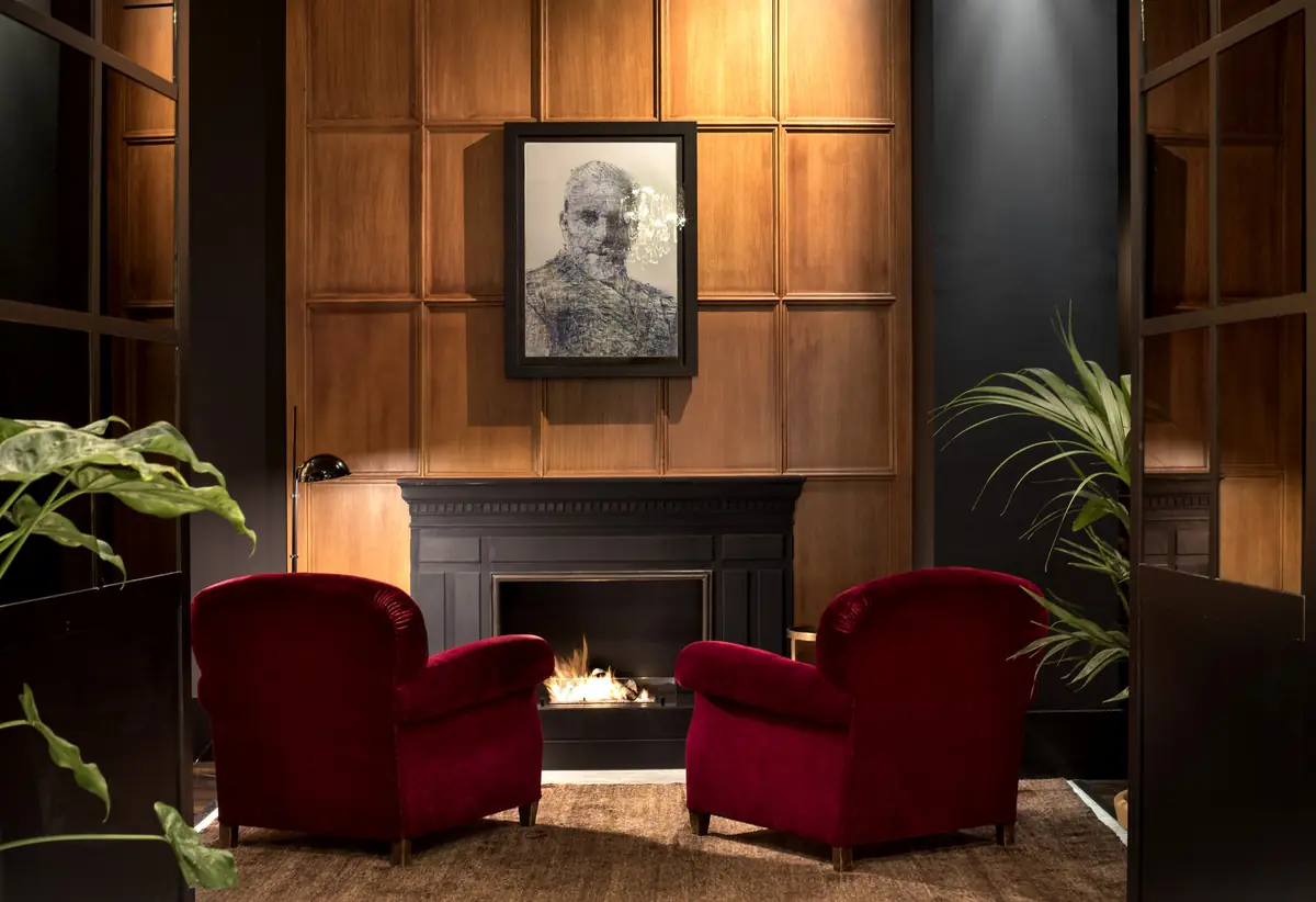 Salón con chimenea, paneles de madera oscura, dos butacas de terciopelo rojo y un retrato enmarcado