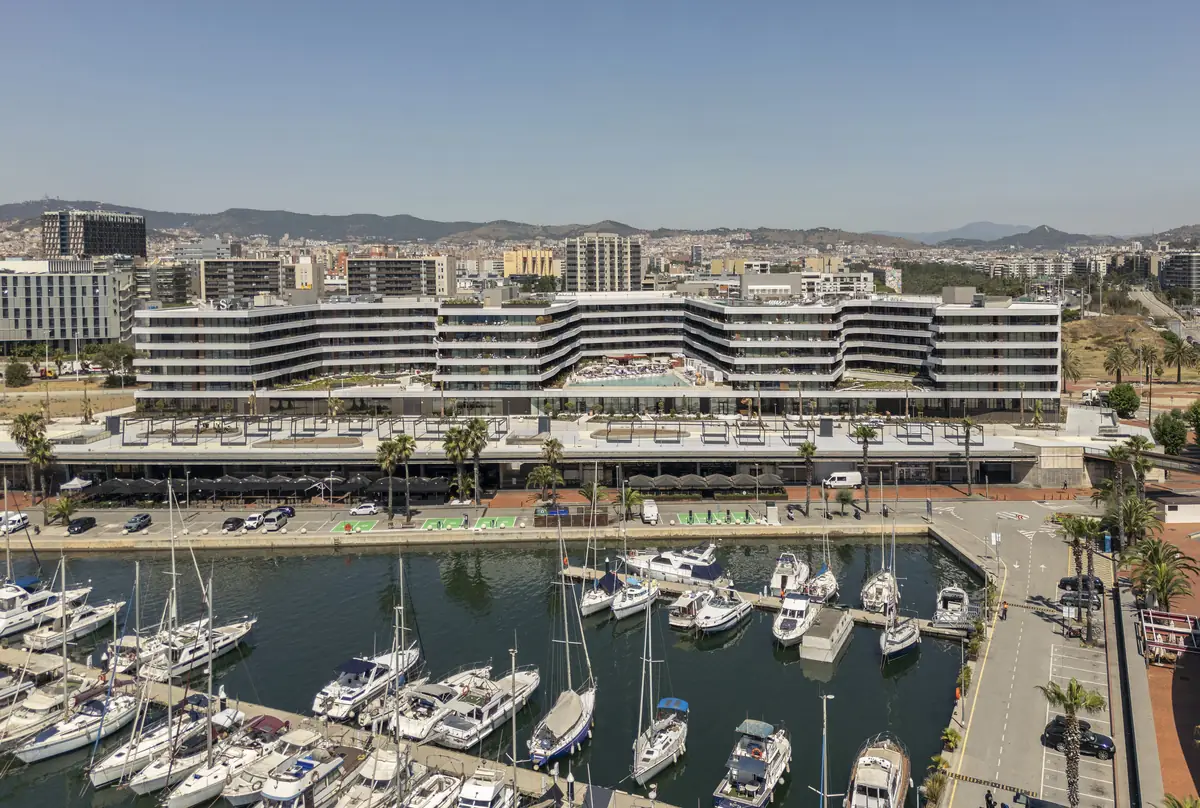Vista aérea de un hotel de planta ondulada frente a un puerto deportivo, con la ciudad y las montañas al fondo