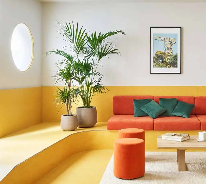 Zona común con banco corrido amarillo, sofá rojo con cojines verdes, pufs naranjas y una palmera en maceta