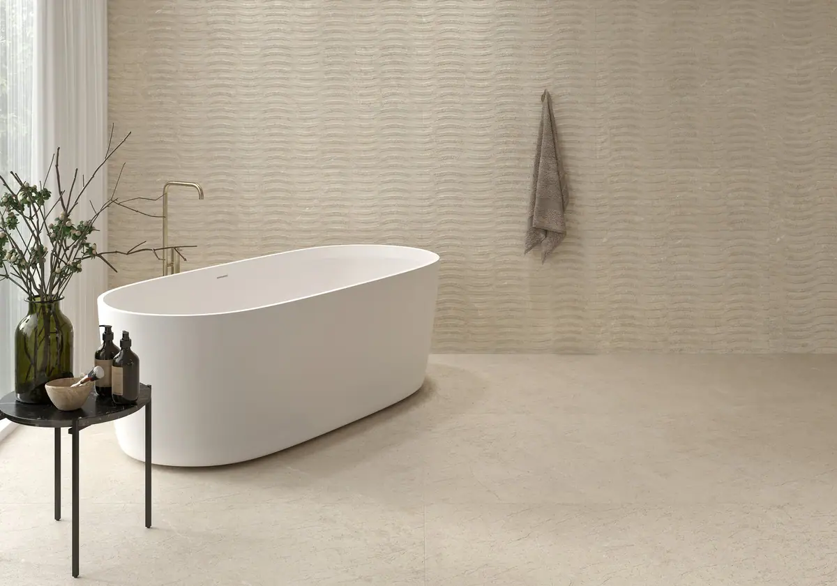 Baño con bañera exenta blanca, pared con relieve ondulado y pavimento de porcelánico efecto piedra en tono claro