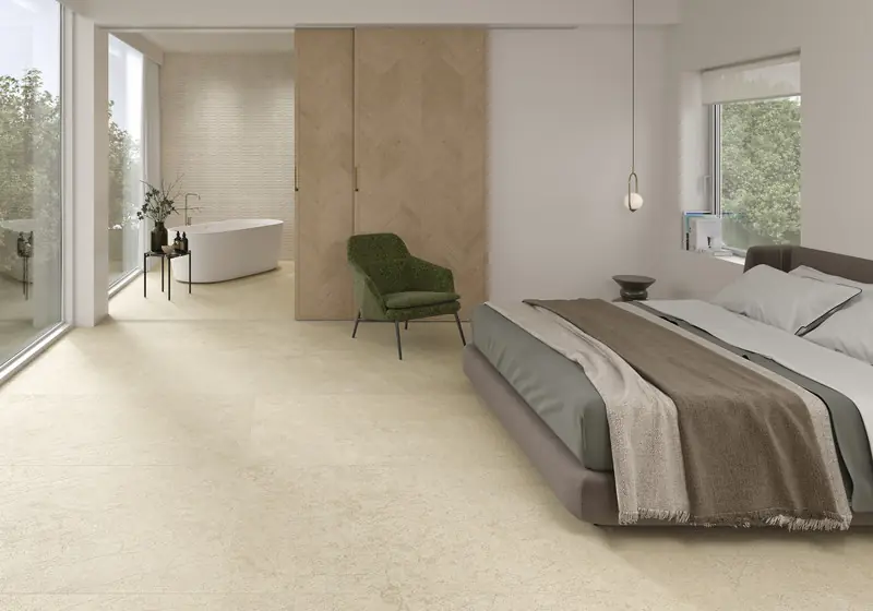 Dormitorio con pavimento de porcelánico efecto piedra clara y, al fondo, un baño con bañera exenta