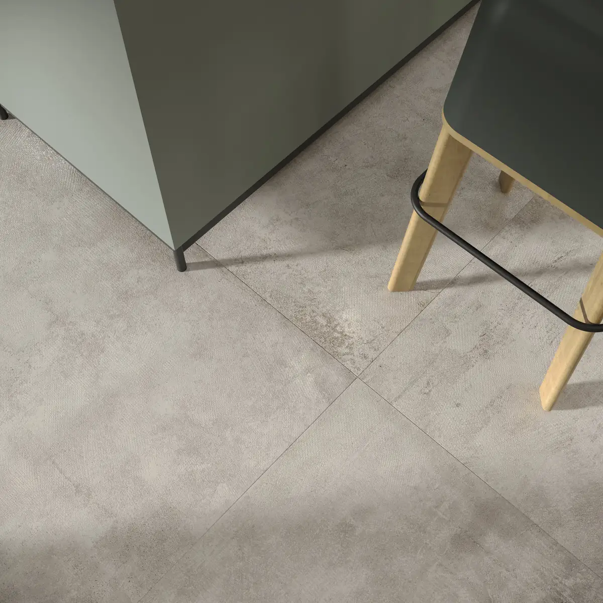 Pavimento de porcelánico efecto cemento gris en gran formato bajo un mueble de cocina y un taburete de madera
