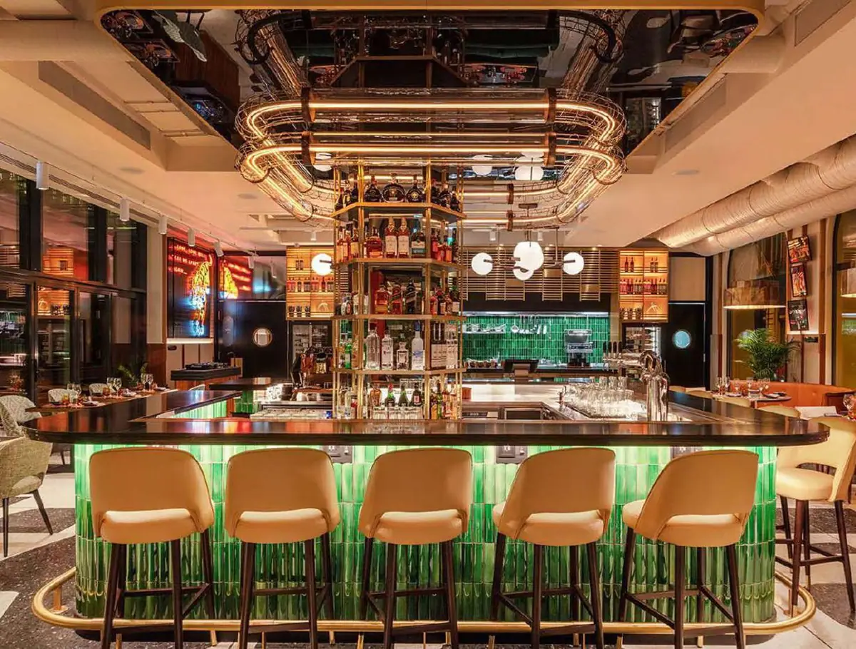 Barra de bar con taburetes tapizados en tono crema y frente de azulejos verdes iluminados