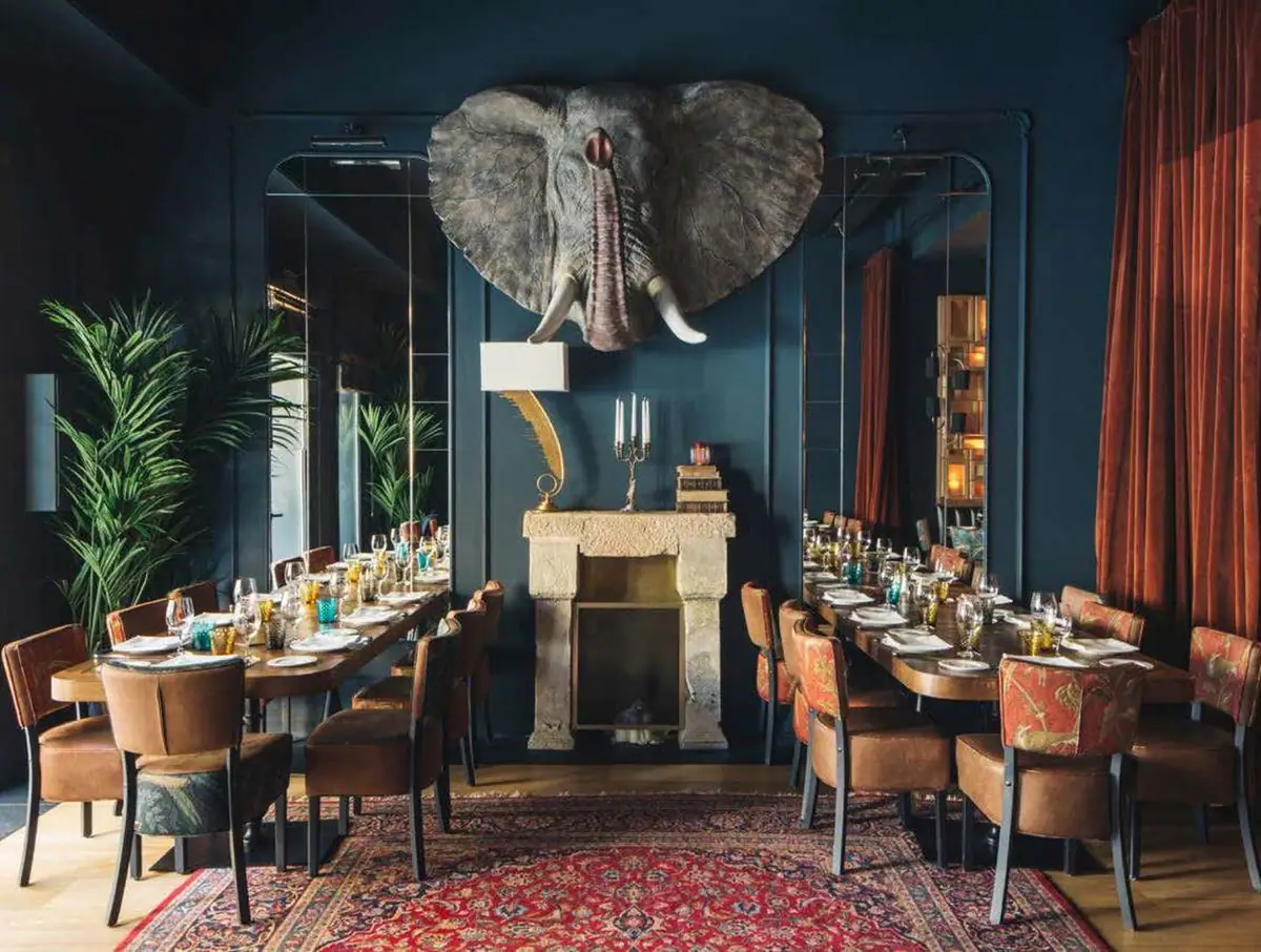 Comedor de paredes azul oscuro con cabeza de elefante sobre la chimenea, sillas tapizadas y cortinas de terciopelo teja