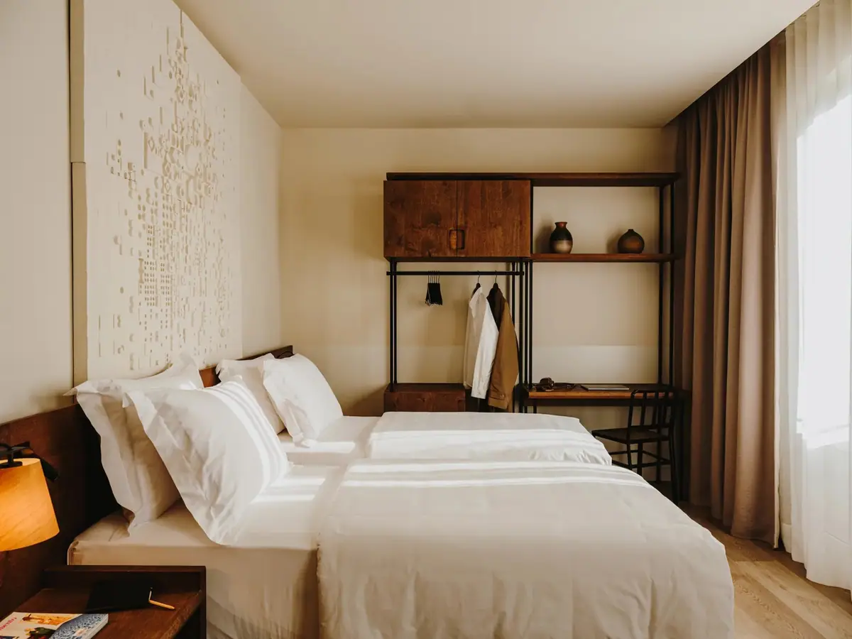 Habitación con dos camas de ropa blanca, cortinas en tono topo, visillo y mueble abierto de madera