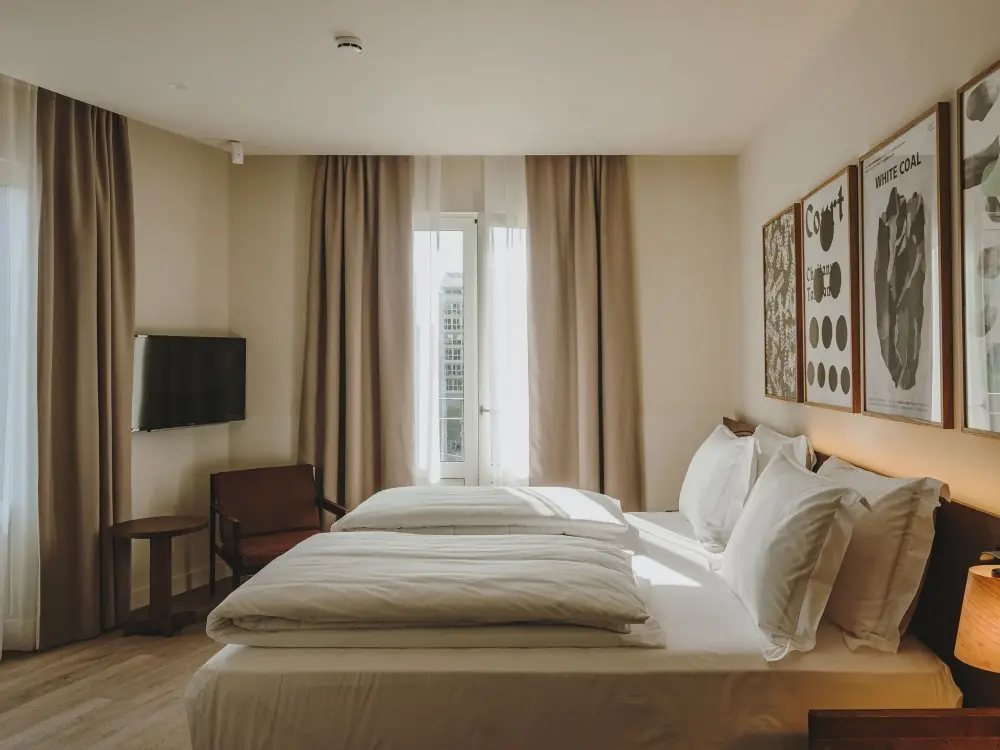 Habitación de hotel con cortinas en tono topo y visillos blancos, cama de matrimonio y láminas enmarcadas