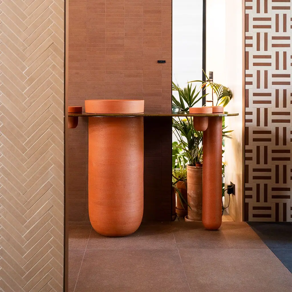 Lavabo de cerámica color terracota sobre una gran columna, con tablero oscuro y una segunda columna más estrecha, ante una pared de ladrillo