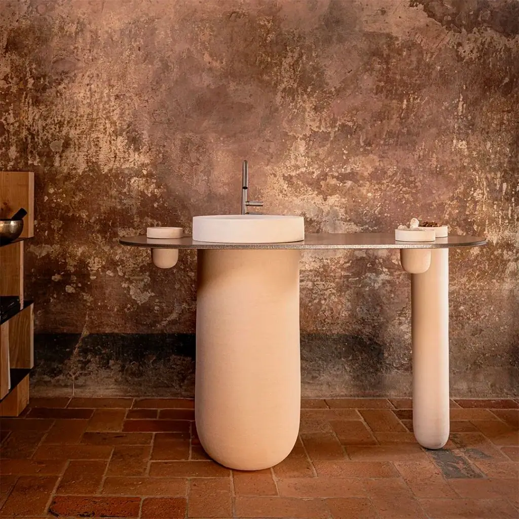 Lavabo blanco sobre columna cerámica de color arena, con tablero metálico y contenedores laterales ante un muro antiguo
