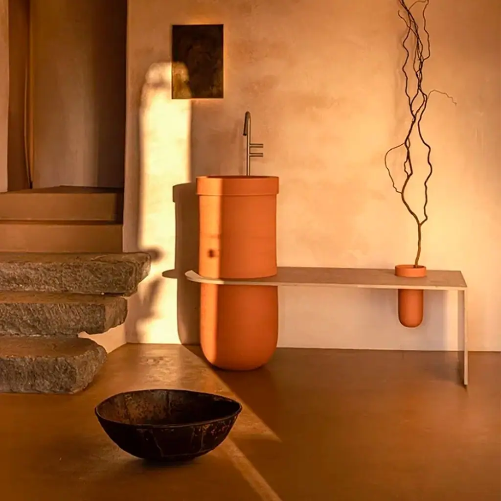 Lavabo cilíndrico de cerámica terracota con un banco de aluminio que sale de él y un pequeño contenedor con ramas secas