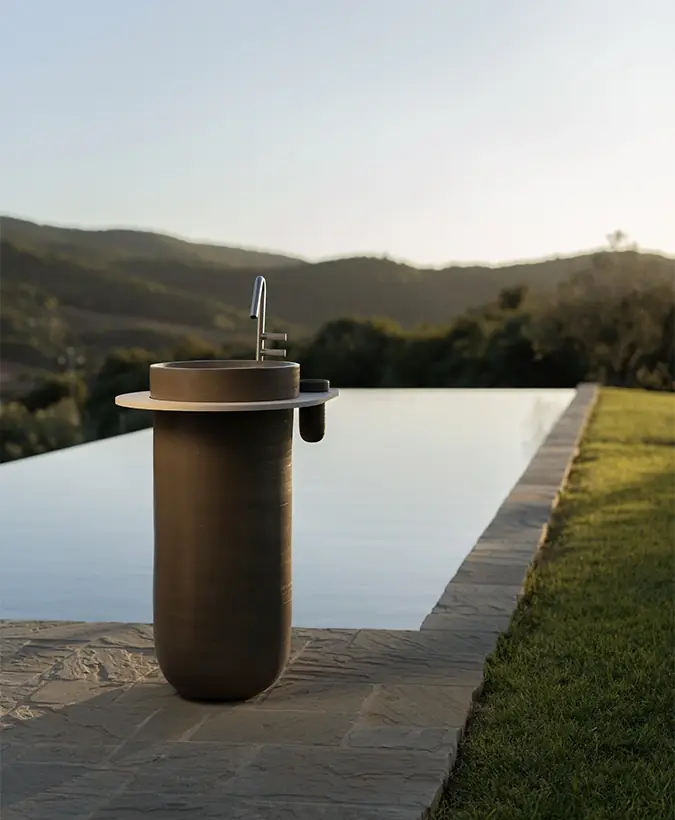 Lavabo de columna de cerámica oscura con tablero redondo junto a una piscina desbordante y colinas al fondo