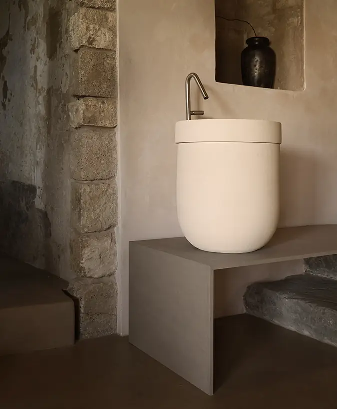 Lavabo alto de cerámica clara sobre un banco bajo, con grifería de acero, junto a un muro de piedra