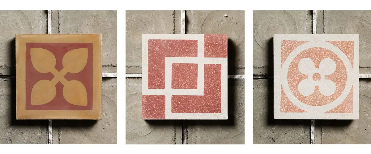 Tres baldosas de cemento decoradas apoyadas sobre una superficie gris: una cementina con hojas amarillas y dos graniglie rojas y blancas