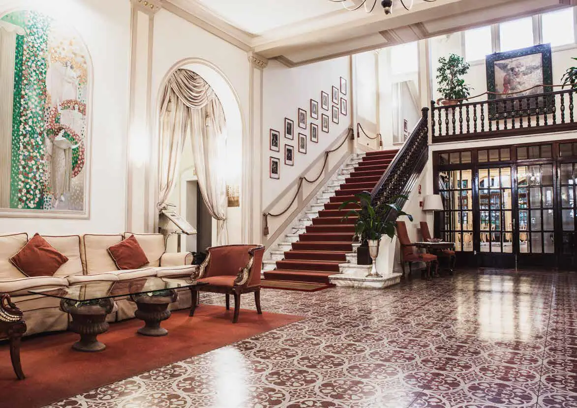 Recepción de un hotel con escalera, sofás y un pavimento de baldosas decoradas en granate y blanco