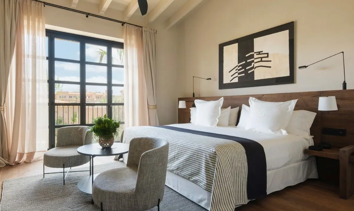 Habitación de hotel con cortinas y visillos de lino claro, butacas tapizadas en gris y colcha de rayas.