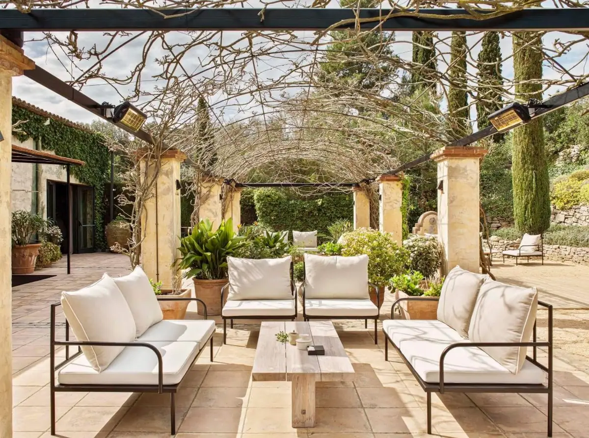 Terraza bajo una pérgola con sofás y butacas exteriores con cojines blancos y mesa de madera.