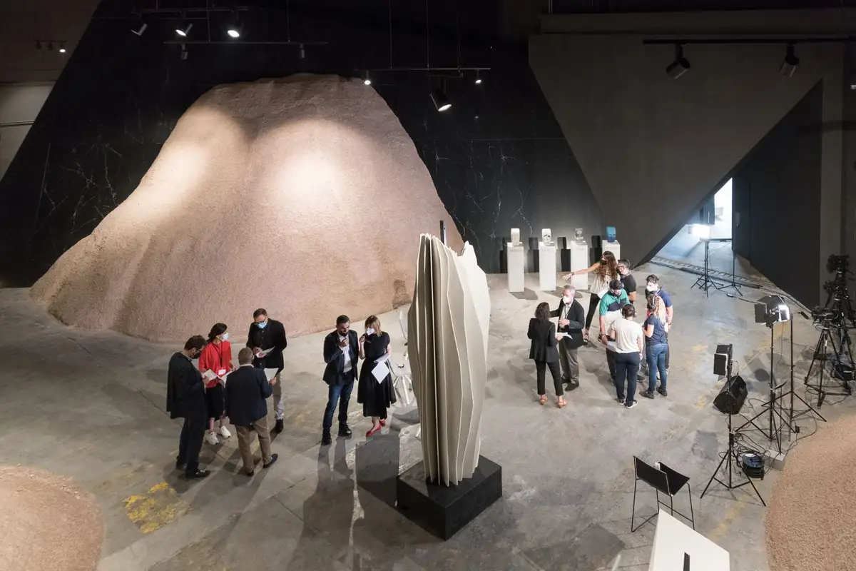 Vista cenital del showroom de COMPAC con grupos de personas conversando junto a una escultura blanca plegada, montículos de material granulado y focos de rodaje.