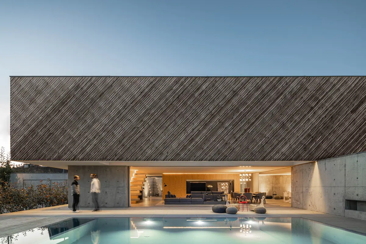 Vivienda con un gran volumen superior revestido de listones de madera en diagonal sobre una planta baja abierta y una piscina