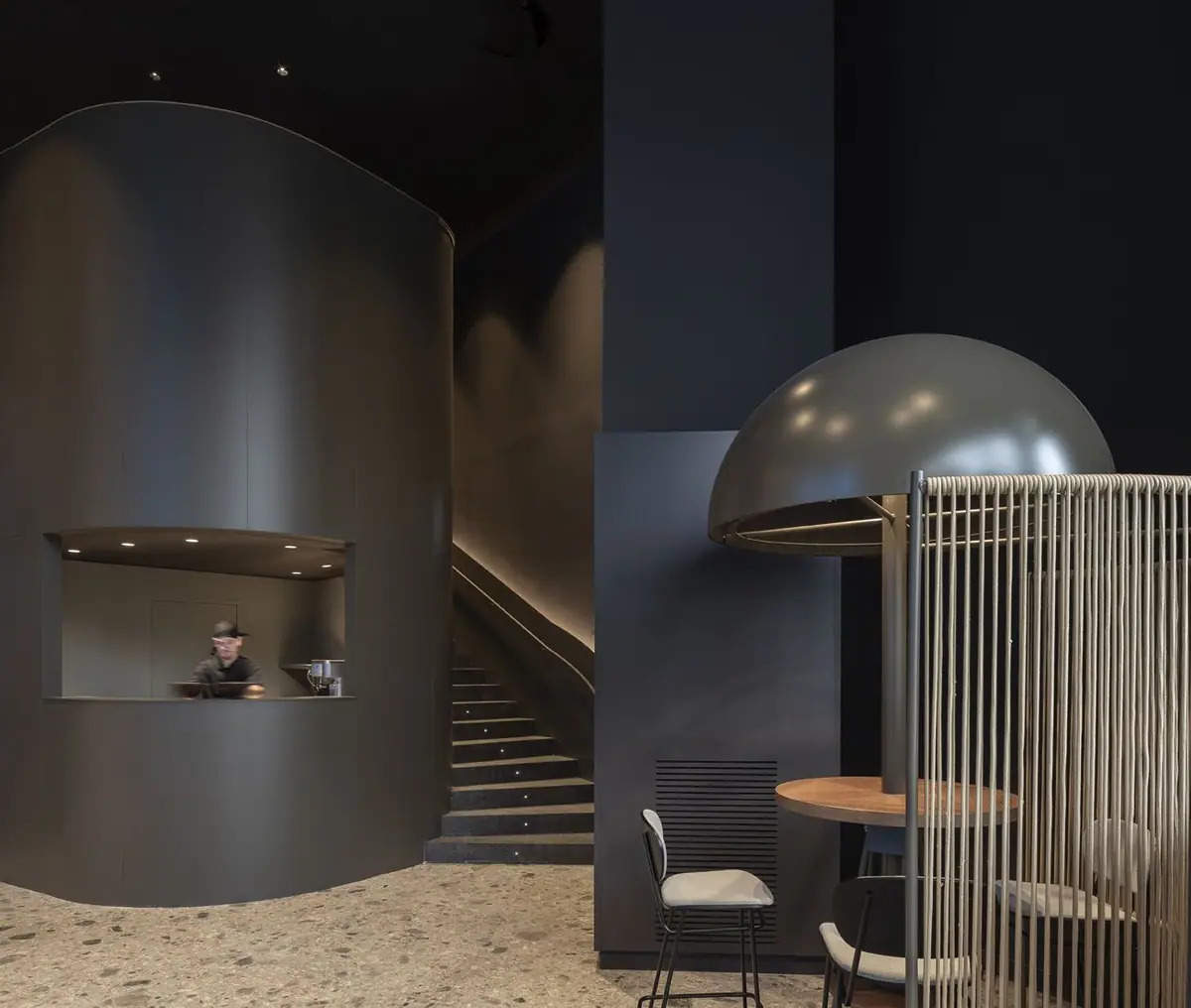 Volumen curvo oscuro con una ventana de servicio, escalera al fondo y mesa alta bajo una cúpula metálica rodeada de cuerdas