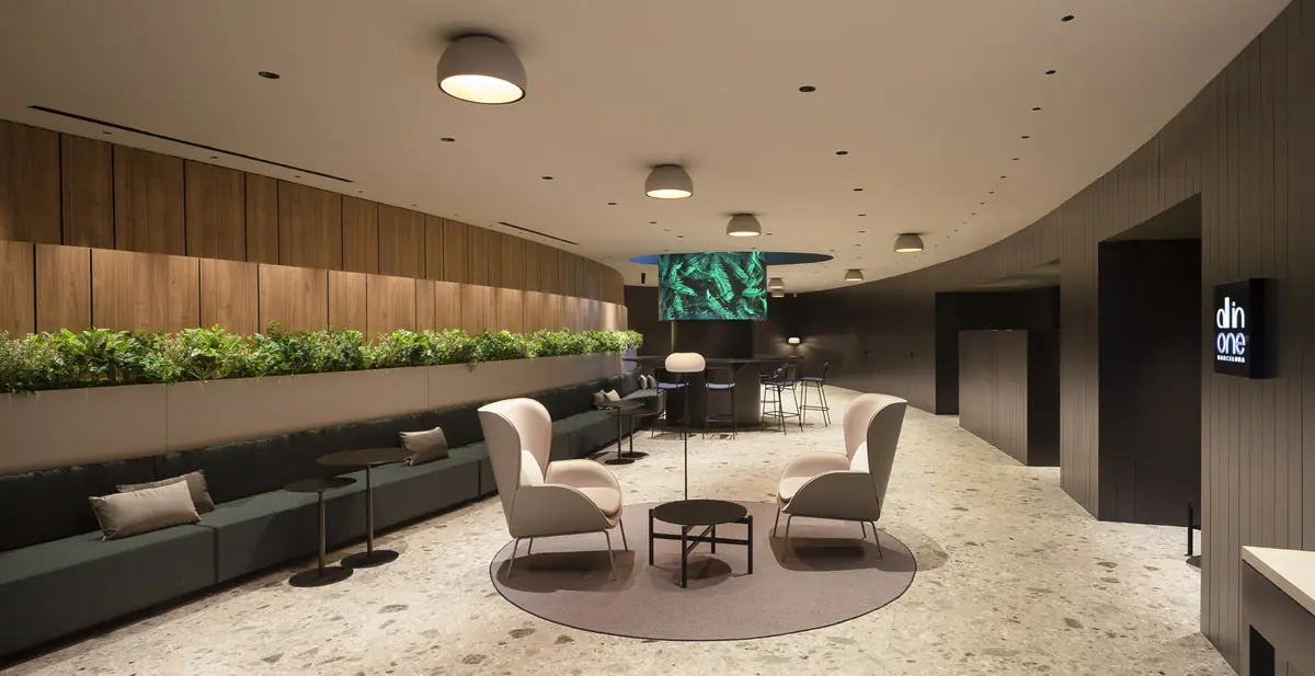 Zona de espera con bancada corrida verde, jardinera, sillones claros y revestimiento de listones de madera en una pared curva