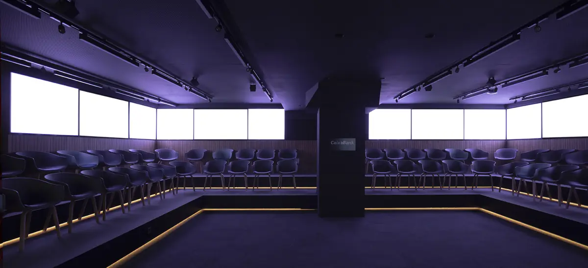 Auditorio con sillas sobre gradas iluminadas y pantallas encendidas en todo el perímetro