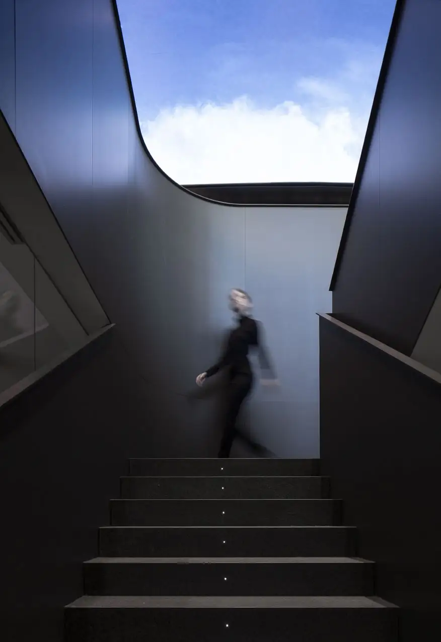Persona subiendo una escalera oscura entre paredes curvas, con una pantalla de cielo en lo alto