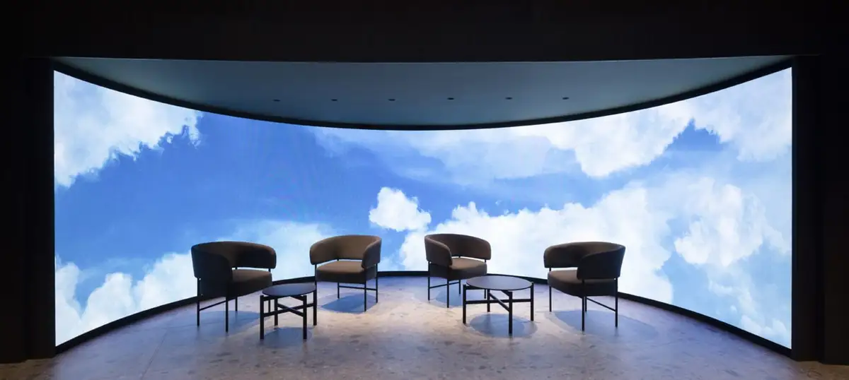 Cuatro butacas y dos mesas bajas frente a una gran pantalla LED curva con imágenes de cielo y nubes