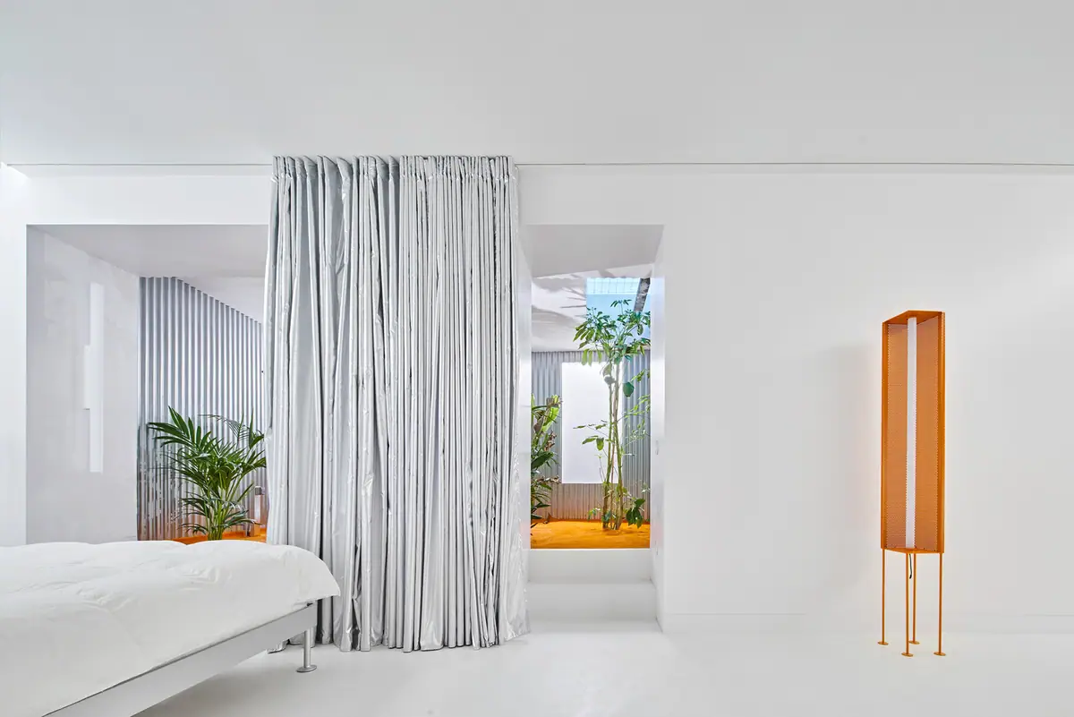 Dormitorio blanco con una cortina plateada y, al fondo, el patio interior con plantas tropicales y césped naranja