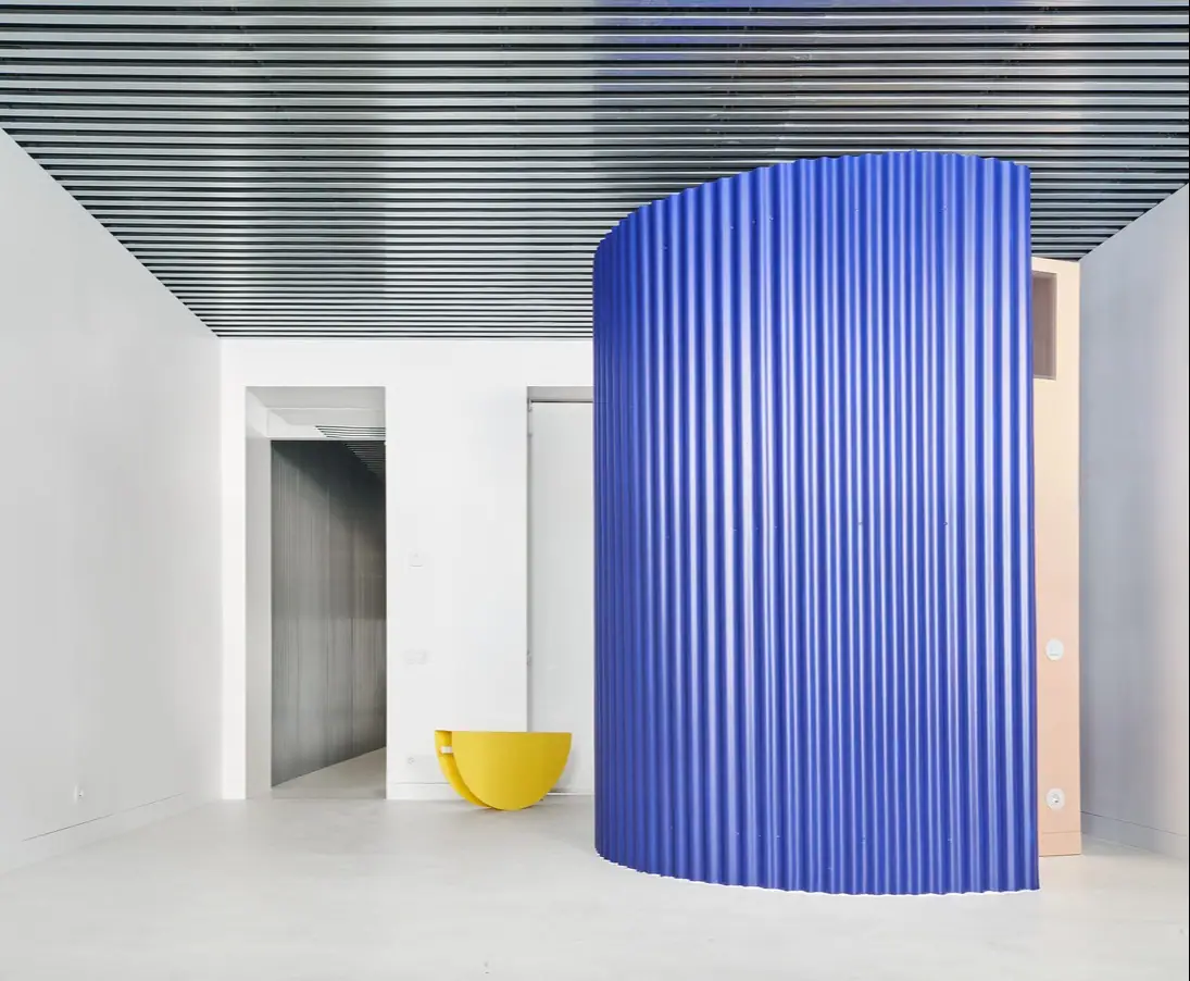 Volumen cilíndrico de chapa ondulada azul bajo un techo de lamas metálicas, con un mueble amarillo junto a la pared