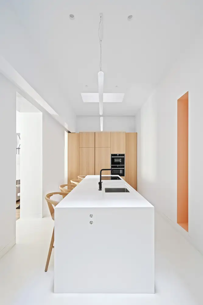 Cocina blanca con isla larga, taburetes de madera, armarios de madera clara y un nicho naranja en la pared