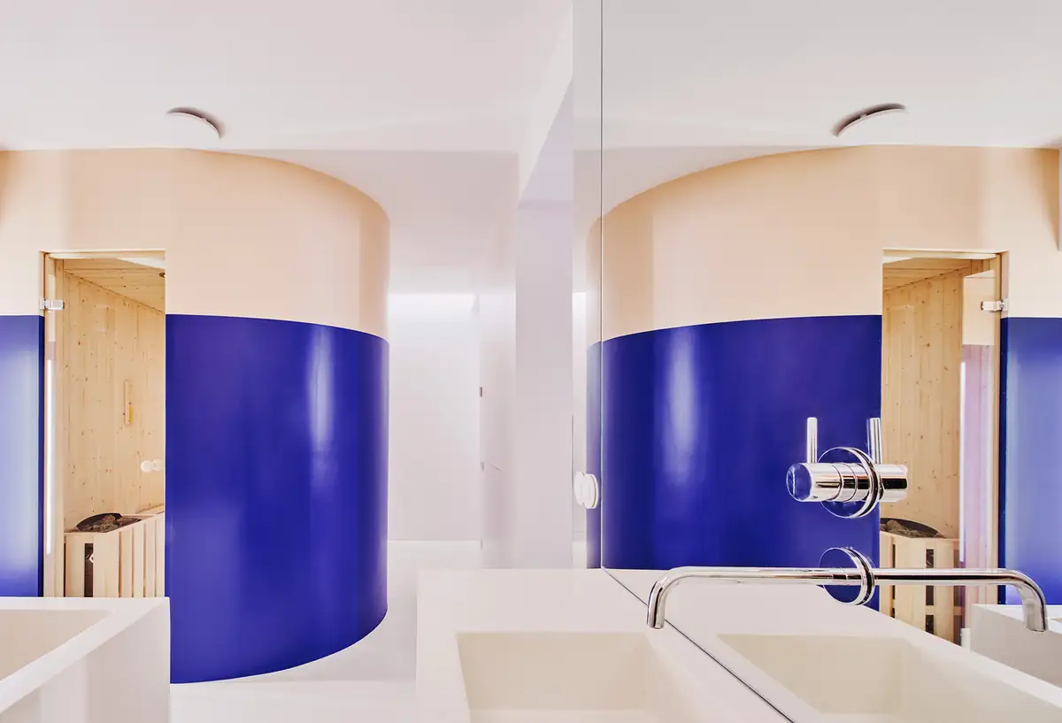 Baño con lavabo blanco frente a un volumen curvo azul y beige que alberga una sauna de madera