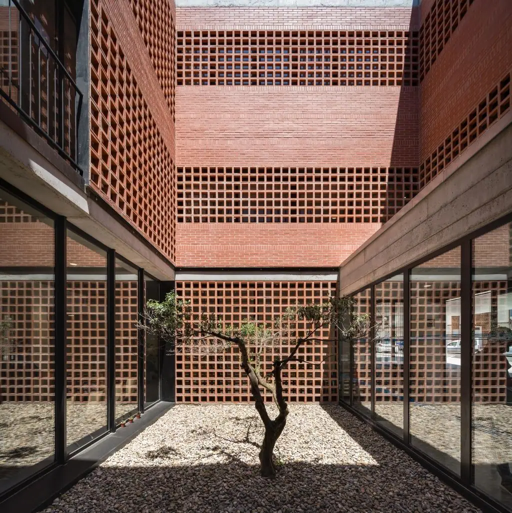 Patio interior con un olivo sobre grava, rodeado de muros de ladrillo y paños de celosía cerámica