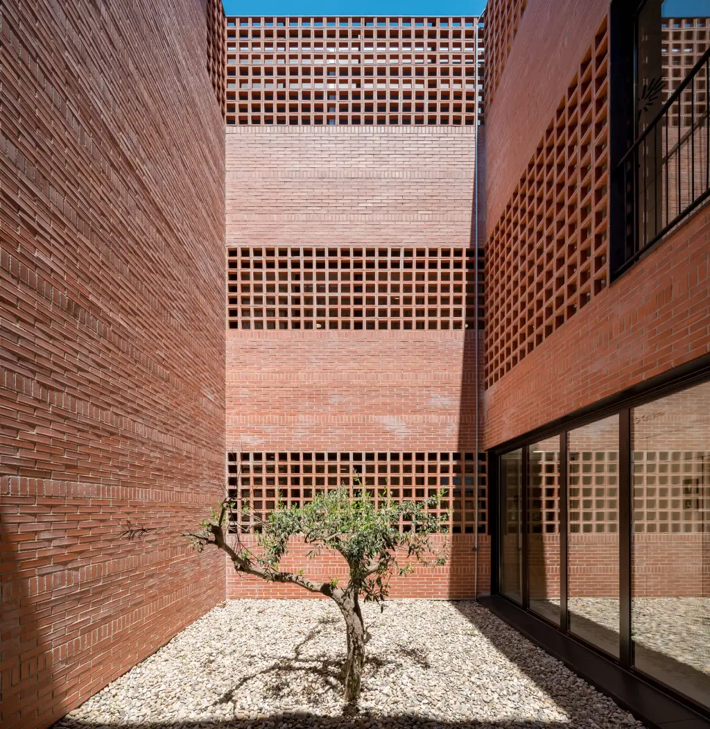 Patio con un olivo entre muros de ladrillo cara vista y bandas horizontales de celosía cerámica