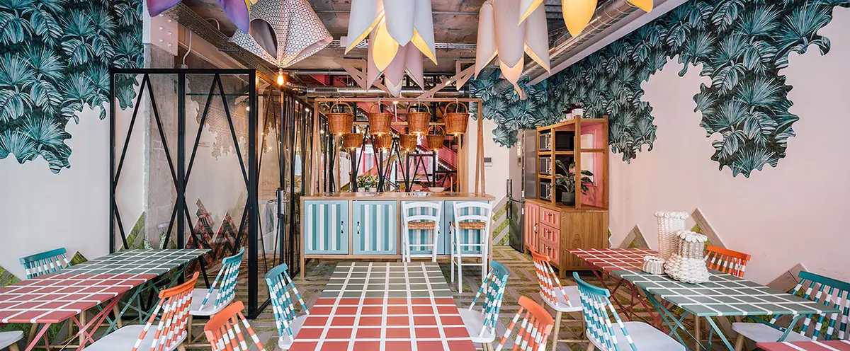Cantina con mesas de cuadros de colores, sillas a rayas, murales de hojas, lámparas en forma de flor y cestas colgadas sobre la barra